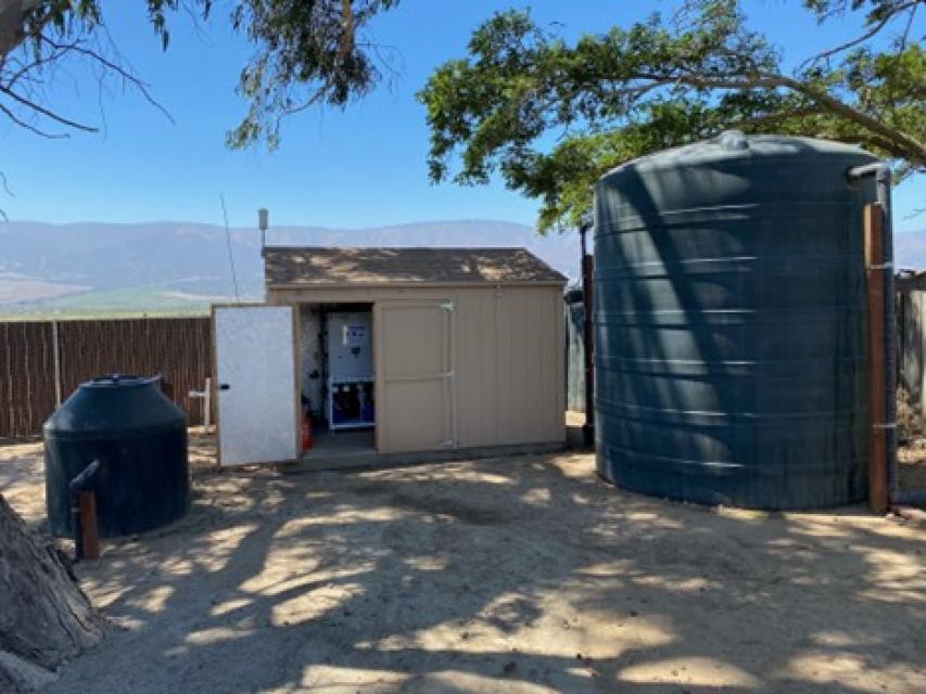 The image shows a large water tank and a shed with water treatment equipment. 